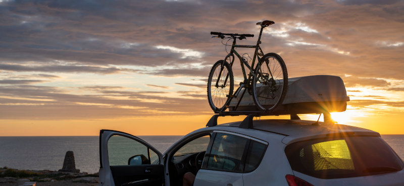 Ein geparktes Auto bei Sonnenuntergang an einer Klippe, mit einem Fahrrad auf dem Dach und einer Dachbox.